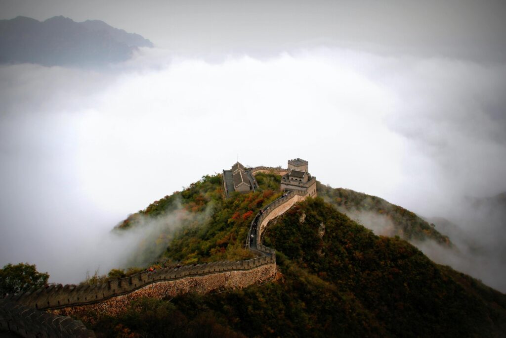 The Great Wall of China winds dramatically along a rugged mountain ridge, seemingly floating above a dense sea of rolling white clouds. Surrounded by lush vegetation and misty peaks, the ancient stone fortifications and watchtowers highlight the structure's imposing scale against the atmospheric landscape.