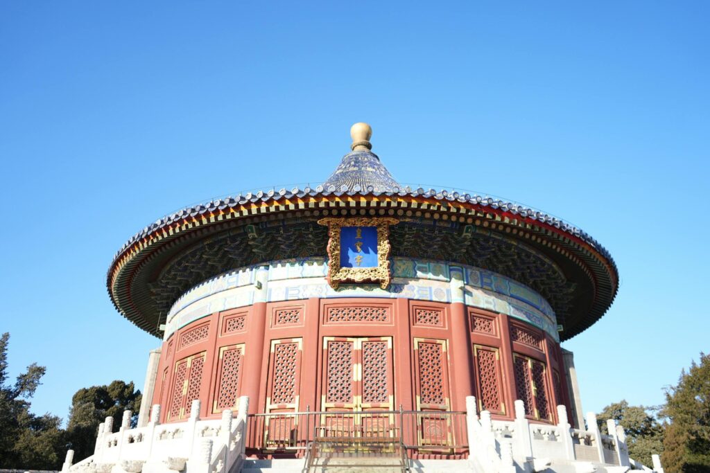 This image showcases the Imperial Vault of Heaven, a circular traditional structure defined by its intricate red lattice walls and a conical, deep blue tiled roof topped with a golden finial. The building stands vividly against a clear azure sky, resting on a white stone base surrounded by carved balustrades that highlight its ornate craftsmanship.