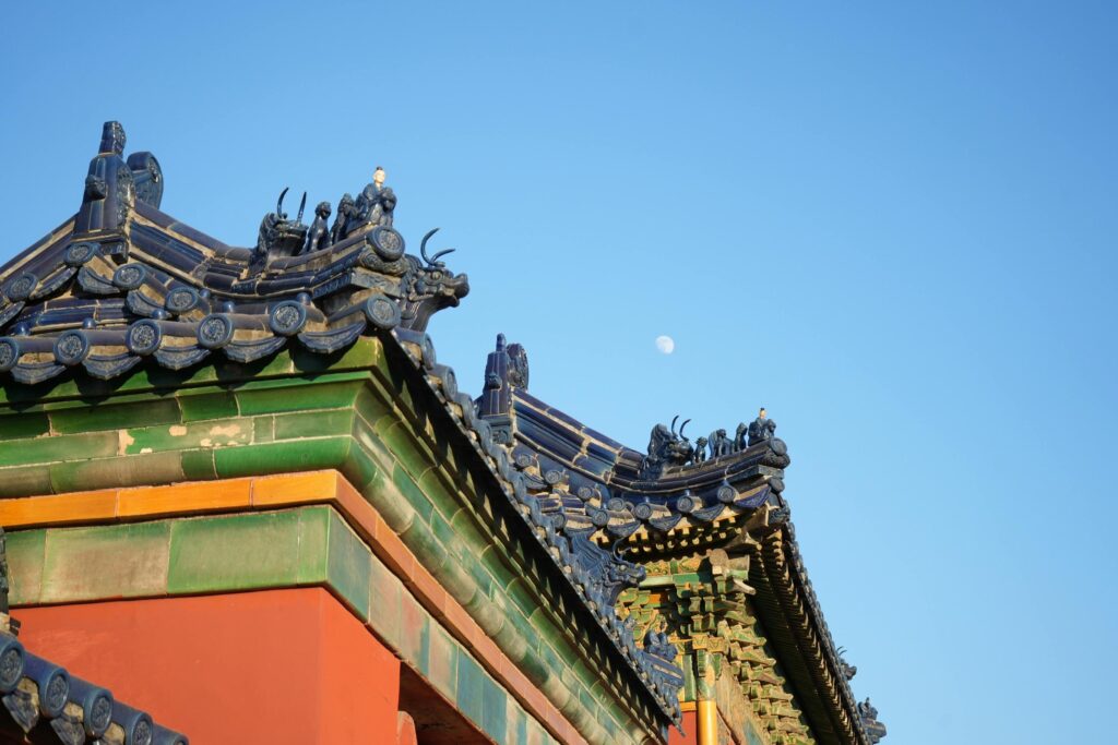 This close-up captures the intricate details of a traditional Chinese roof, featuring deep blue glazed tiles and rows of mythical beast figurines perched along the ridges. Set against a clear blue sky with a faint moon, the vibrant green and blue roofing contrasts boldly with the rich red wall beneath.