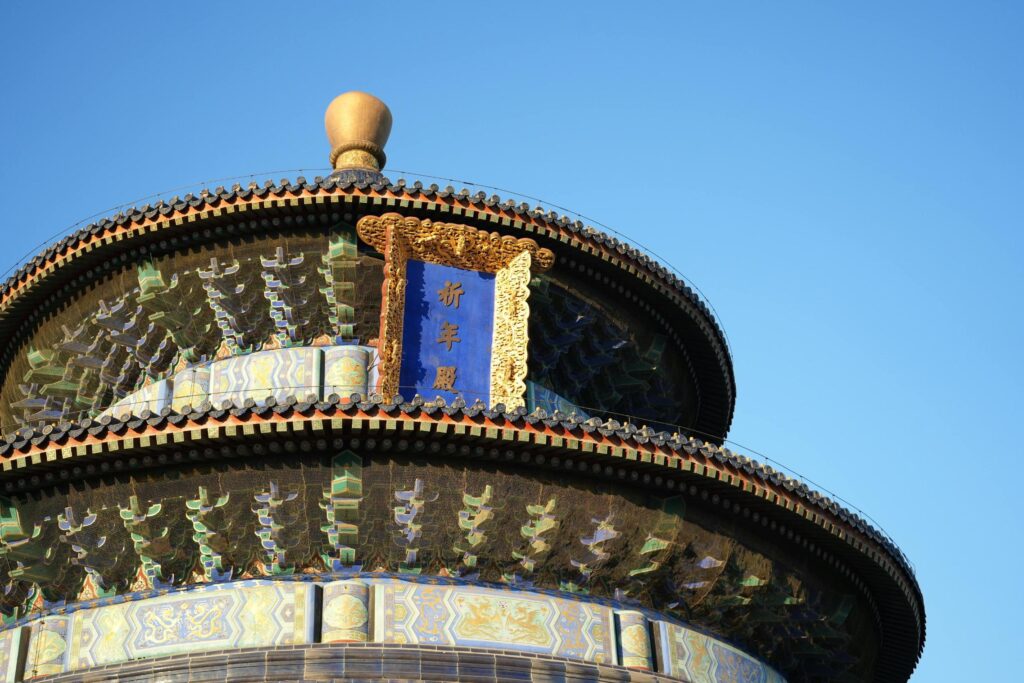 This low-angle shot highlights the intricate upper tiers of the Hall of Prayer for Good Harvests, showcasing vibrant blue roof tiles and complex, multi-colored wooden brackets beneath the eaves. A central blue plaque featuring golden Chinese characters is framed by ornate gold detailing, while a golden finial crowns the circular roof against a bright azure sky.