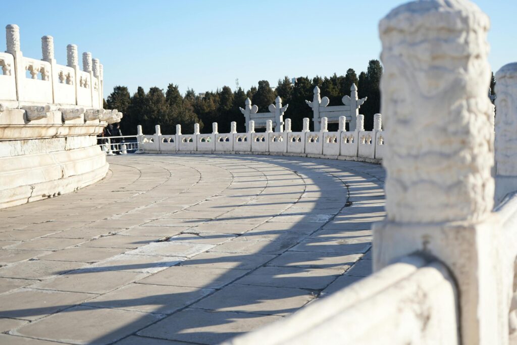 This image depicts a curved, paved terrace lined with white marble balustrades, where intricately carved posts cast rhythmic shadows across the stone floor. In the background, the walkway follows the contour of a circular stone base, bordered by a line of dark green trees under a bright, clear sky.