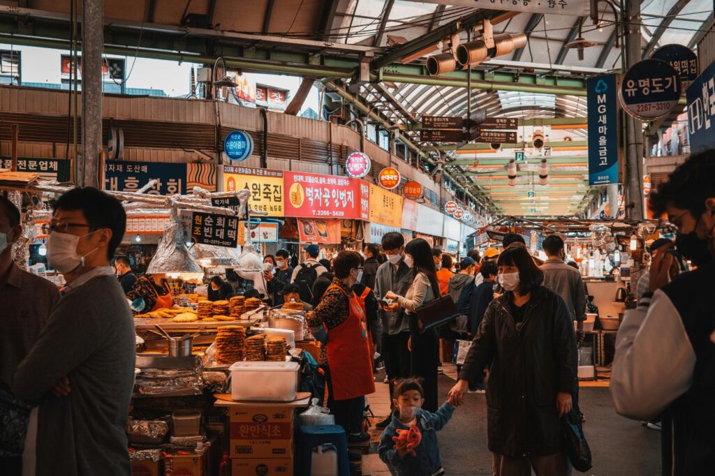 A busy indoor market comes to life with crowds of mask-wearing shoppers navigating through narrow aisles lined with food stalls and bright Korean signage. In the foreground, a young child holding a toy walks hand-in-hand with a woman past vendors displaying stacks of fried goods under the covered roof.