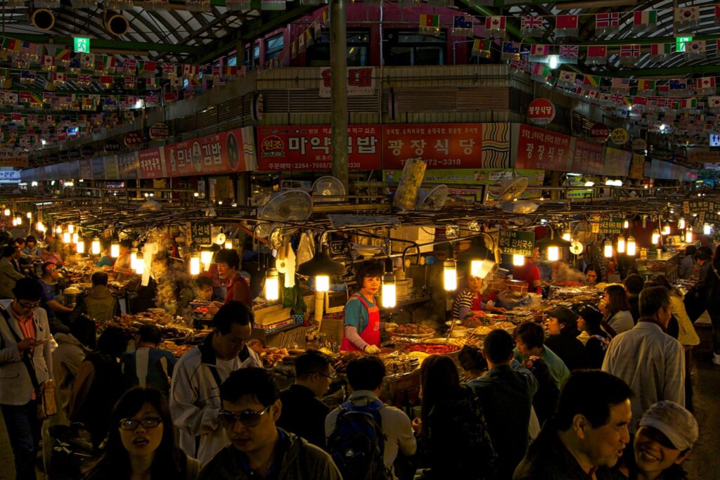 Under the warm glow of numerous hanging lightbulbs, a crowded night market bustles with activity as vendors serve steaming food to customers seated at central stalls. The lively scene features international flags suspended from the ceiling and Korean signage lining the upper levels, framing the dense throng of visitors below.
