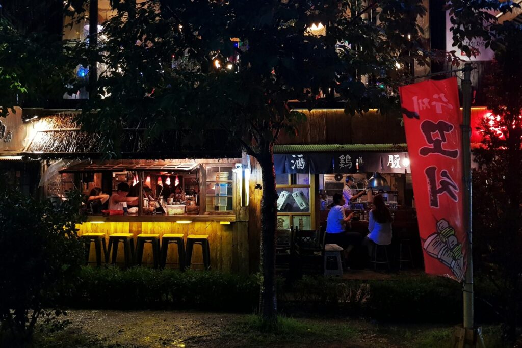 Framed by dark trees at night, a cozy wooden eatery emits a warm glow from its open windows where patrons dine and staff work behind the counter. A bright red vertical banner stands prominently to the right, contrasting with the row of yellow stools lined up beneath the thatched roof overhang on the left.