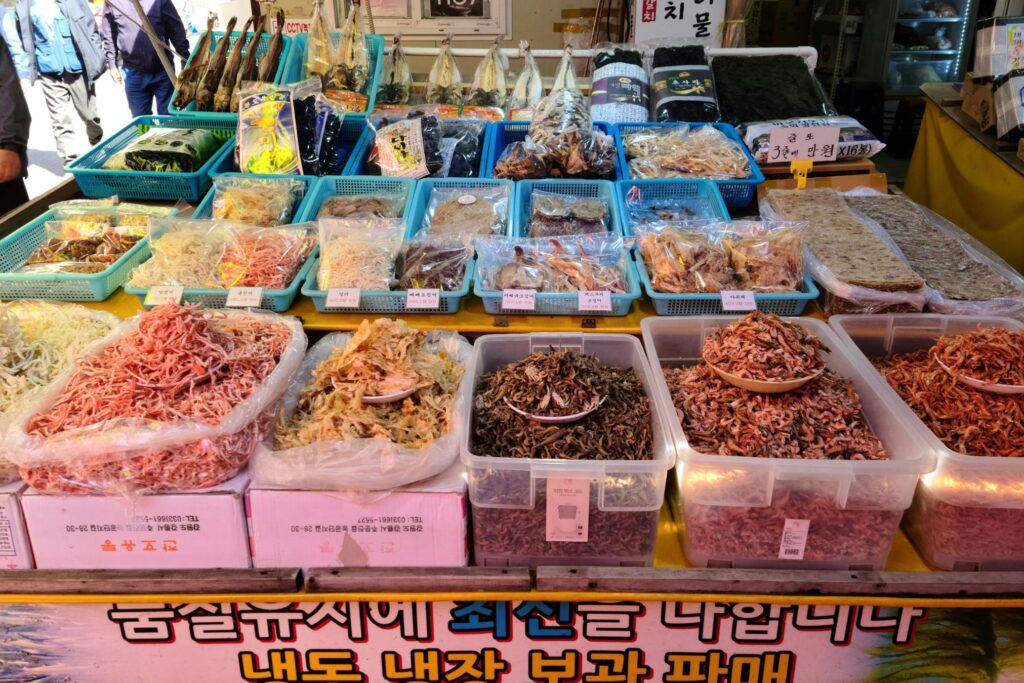 Rows of blue baskets and clear bags display a variety of dried seafood, including whole fish, shredded squid, and seaweed packages, arranged neatly on a market stall. Beneath the upper tiers, large bins and open sacks are filled with bulk quantities of tiny dried shrimp and anchovies, resting above boxes printed with Korean text.
