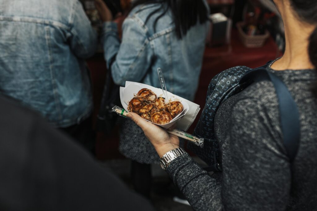 This close-up shot captures a person holding a paper tray filled with golden-brown takoyaki, garnished with savory sauce and bonito flakes. In the background, blurred figures in denim and casual wear suggest a lively, crowded street food environment.