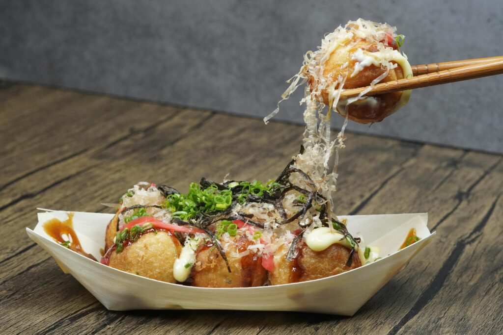 Wooden chopsticks hold a single takoyaki ball aloft, creating a bridge of delicate bonito flakes connecting it to the paper boat below. The dish is presented on a wooden table, generously topped with savory brown sauce, creamy mayonnaise, green seaweed, and pickled ginger.