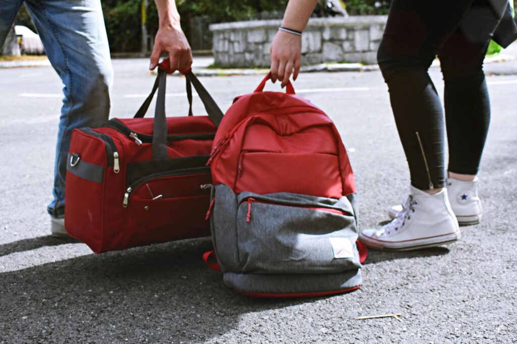 Two travelers, visible only from the waist down, are shown on an asphalt road handling their luggage. One person holds a red duffel bag while the other, wearing white high-top sneakers, grips the top of a red and grey backpack.