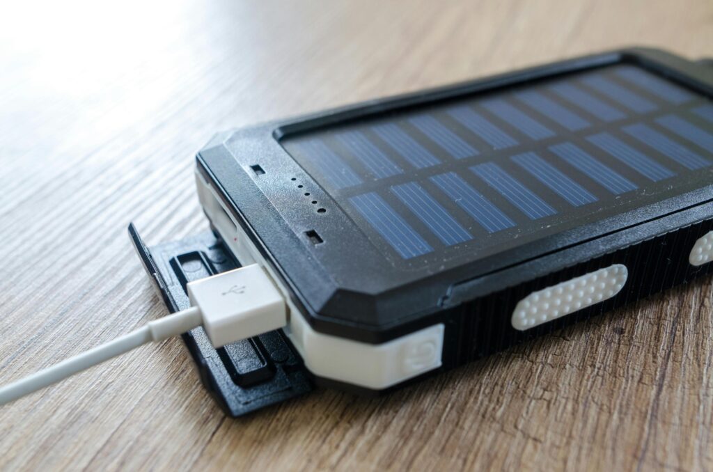 A rugged black power bank with an integrated solar panel rests on a light wooden surface. A white USB cable is plugged into the side of the device, indicating it is ready to charge or transfer power.