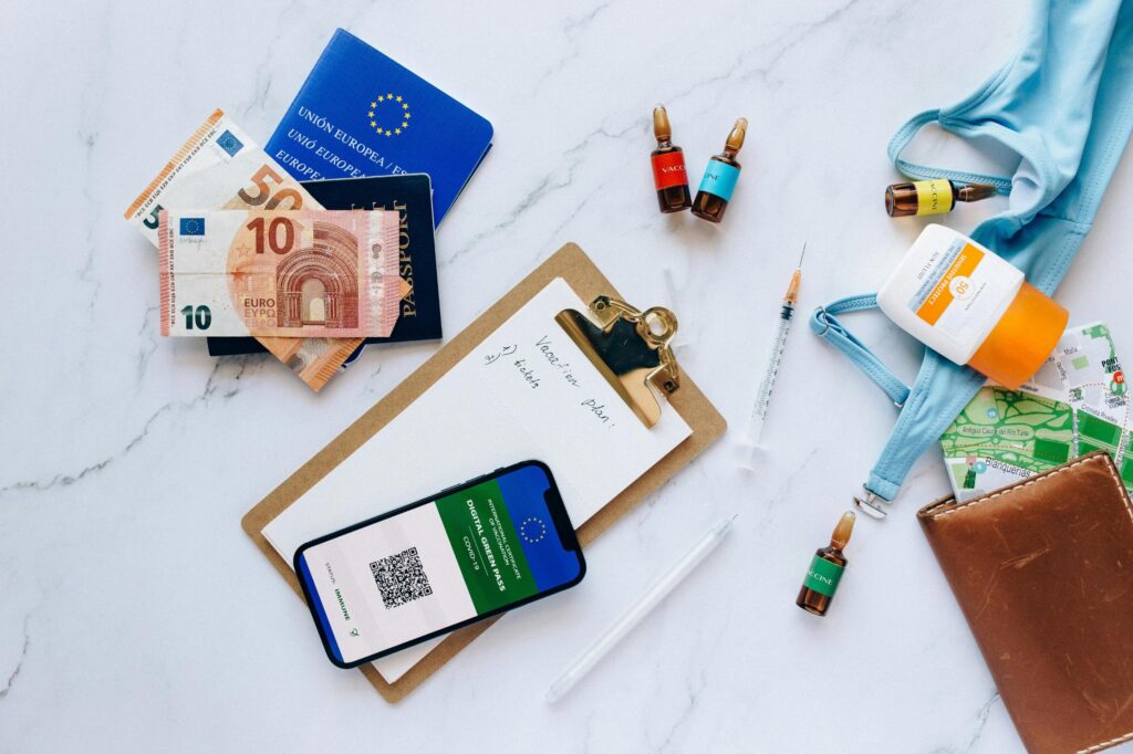 A flat-lay composition on a white marble background displays travel essentials, including a European Union passport, Euro banknotes, and a smartphone showing a COVID-19 Digital Green Pass. Surrounding the documents are vacation items like a blue swimsuit, sunscreen, and vaccine vials, highlighting the specific preparations needed for travel during the pandemic.