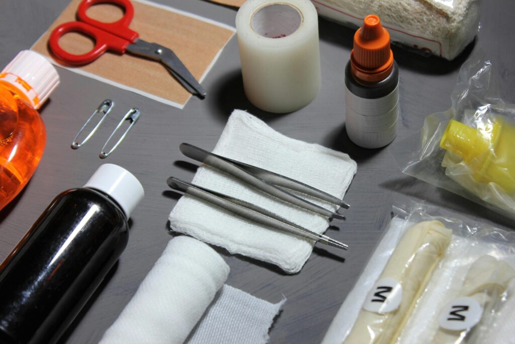 A neatly organized collection of first aid supplies is displayed on a grey surface, featuring essential items such as tweezers resting on a white gauze pad, rolled bandages, and several bottles of antiseptic solutions. The kit also includes red-handled scissors, safety pins, adhesive tape, and packaged gloves, suggesting a comprehensive set of tools for treating minor injuries.