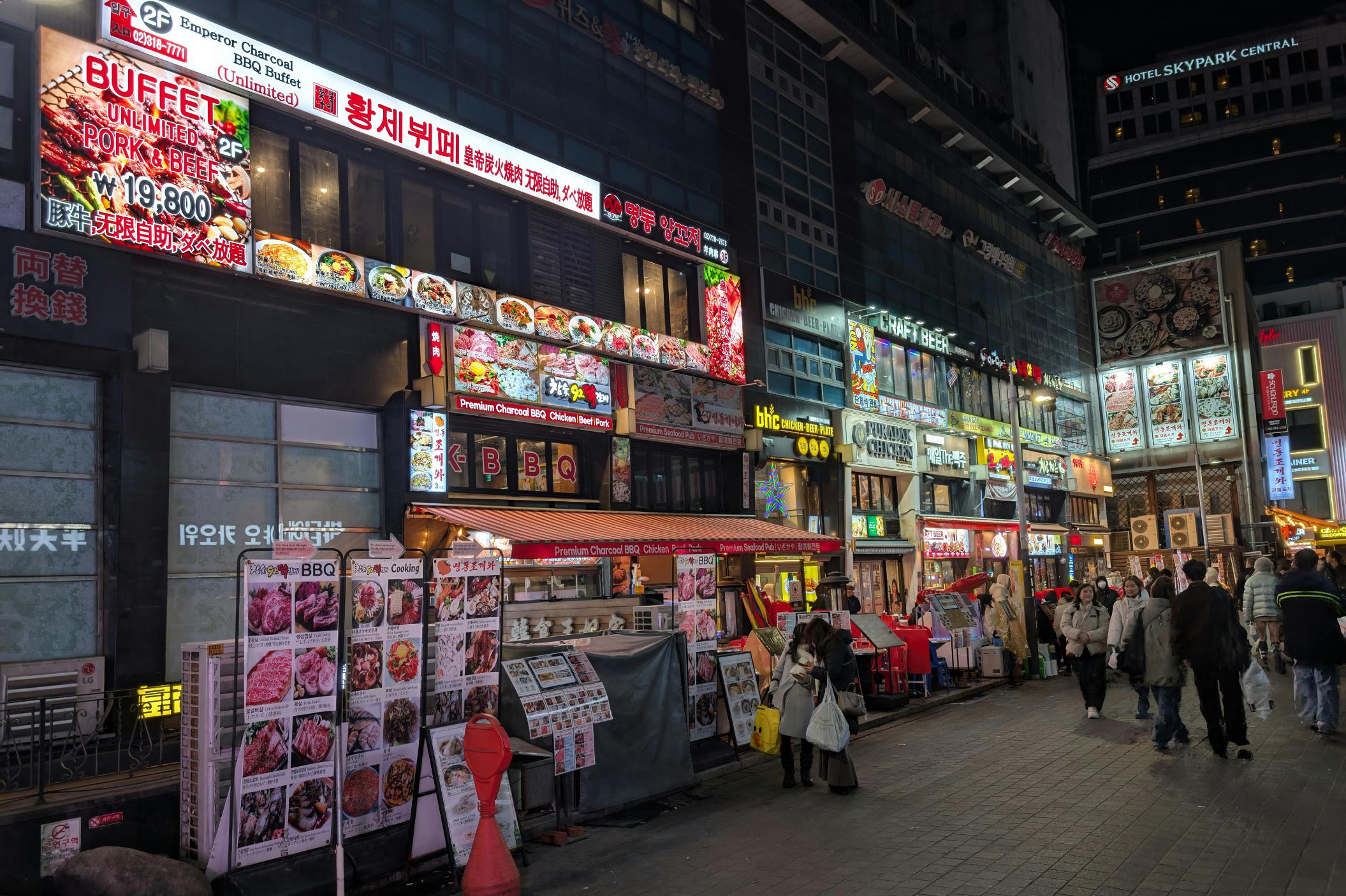 A bustling city street at night is illuminated by vibrant signage, including a prominent advertisement for an "Emperor Charcoal BBQ Buffet". Pedestrians stroll along the sidewalk past rows of menu stands displaying colorful photographs of various meat dishes outside the restaurants.