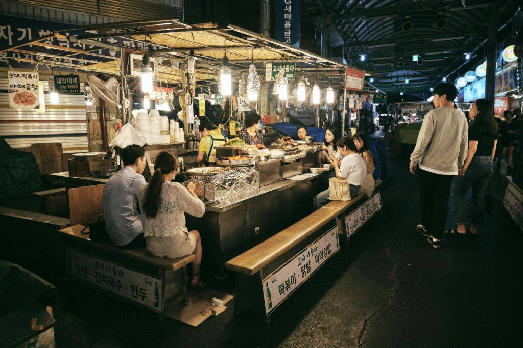 Under the warm glow of hanging light bulbs, customers sit on wooden benches at a busy street food stall, enjoying a meal directly at the counter. The lively market scene features vendors preparing food behind mounds of ingredients, while pedestrians stroll through the dimly lit covered alleyway lined with Korean signage.