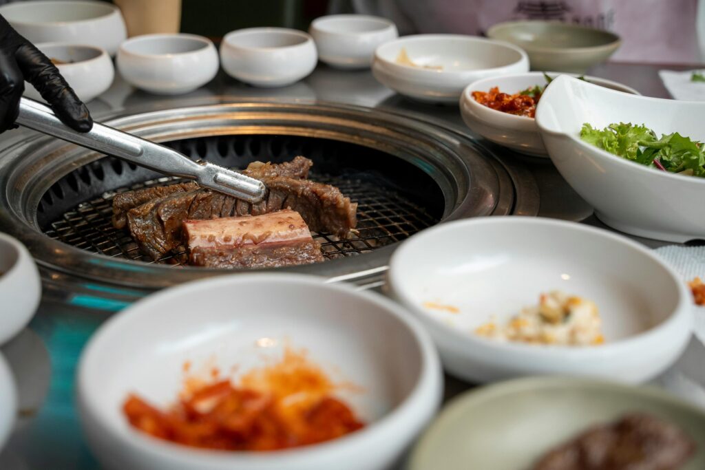 A gloved hand uses metal tongs to grill marinated beef short ribs on a circular grate embedded in the center of a dining table. The main dish is surrounded by an assortment of traditional Korean side dishes, or banchan, served in small white bowls, including kimchi and fresh salad.
