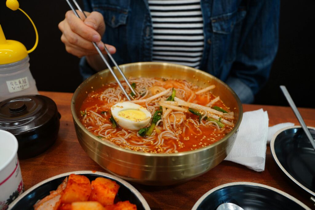 A diner in a denim jacket and striped shirt uses chopsticks to eat a bowl of cold noodle soup Naengmyeon served in a brass bowl with a hard-boiled egg and radish garnish. The table is set with traditional accompaniments, including a dish of spicy radish kimchi Kkakdugi, a bottle of vinegar, and metal side dishes.