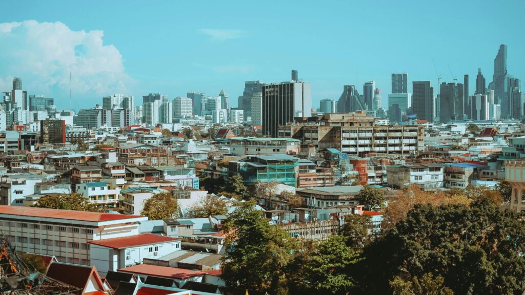 This image captures an expansive view of Bangkok’s skyline, showcasing a dense contrast between low-rise residential neighborhoods and towering modern skyscrapers. Under a bright blue sky, the cityscape blends traditional architecture and greenery with the sleek, metallic silhouettes of the city's financial district.