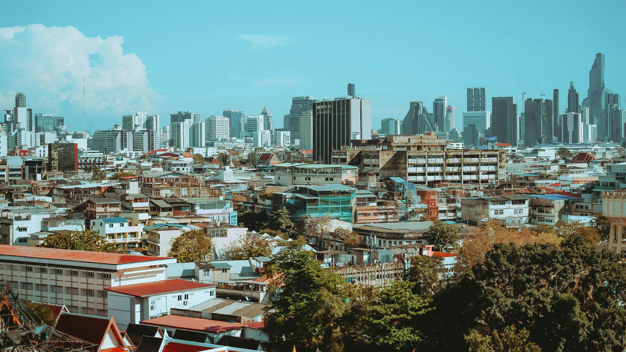 This image captures an expansive view of Bangkok’s skyline, showcasing a dense contrast between low-rise residential neighborhoods and towering modern skyscrapers. Under a bright blue sky, the cityscape blends traditional architecture and greenery with the sleek, metallic silhouettes of the city's financial district.
