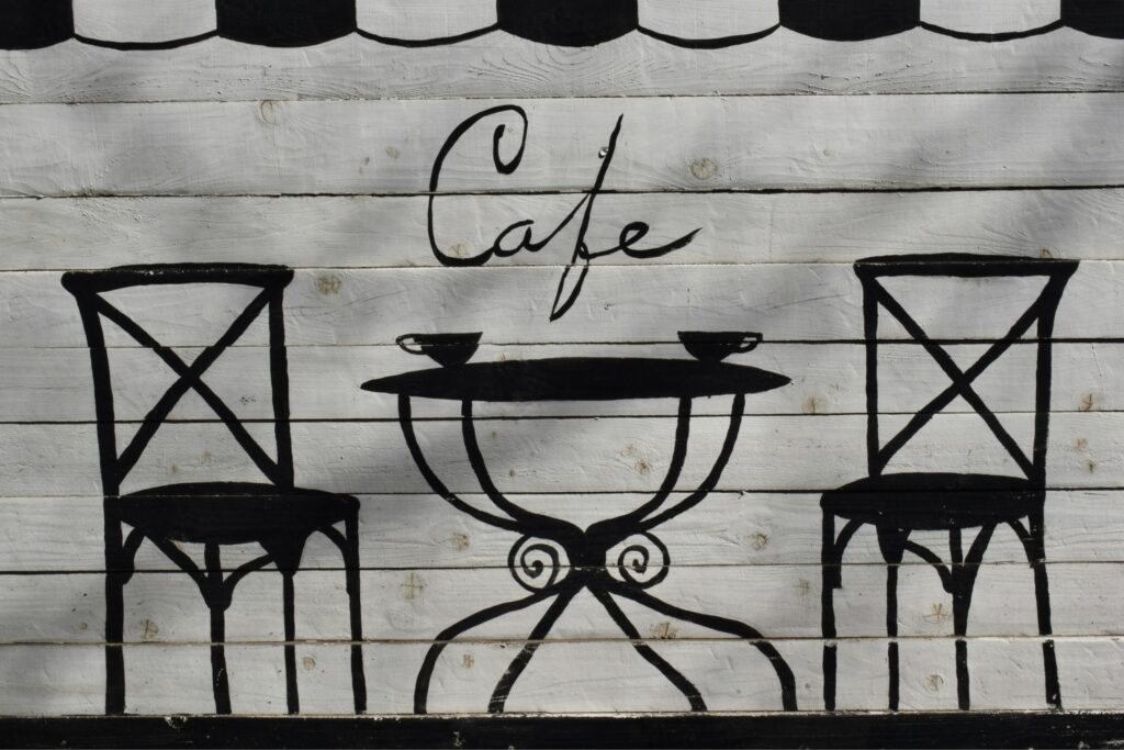 This image features a charming, hand-painted mural on a white wooden plank wall depicting a minimalist cafe scene. Two black bistro chairs flank a small table with two cups, set beneath elegant script that reads "Cafe”.