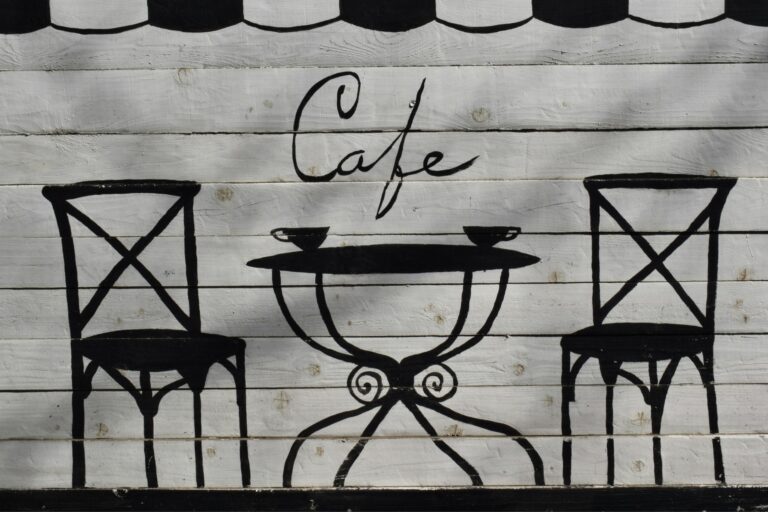 This image features a charming, hand-painted mural on a white wooden plank wall depicting a minimalist cafe scene. Two black bistro chairs flank a small table with two cups, set beneath elegant script that reads "Cafe”.