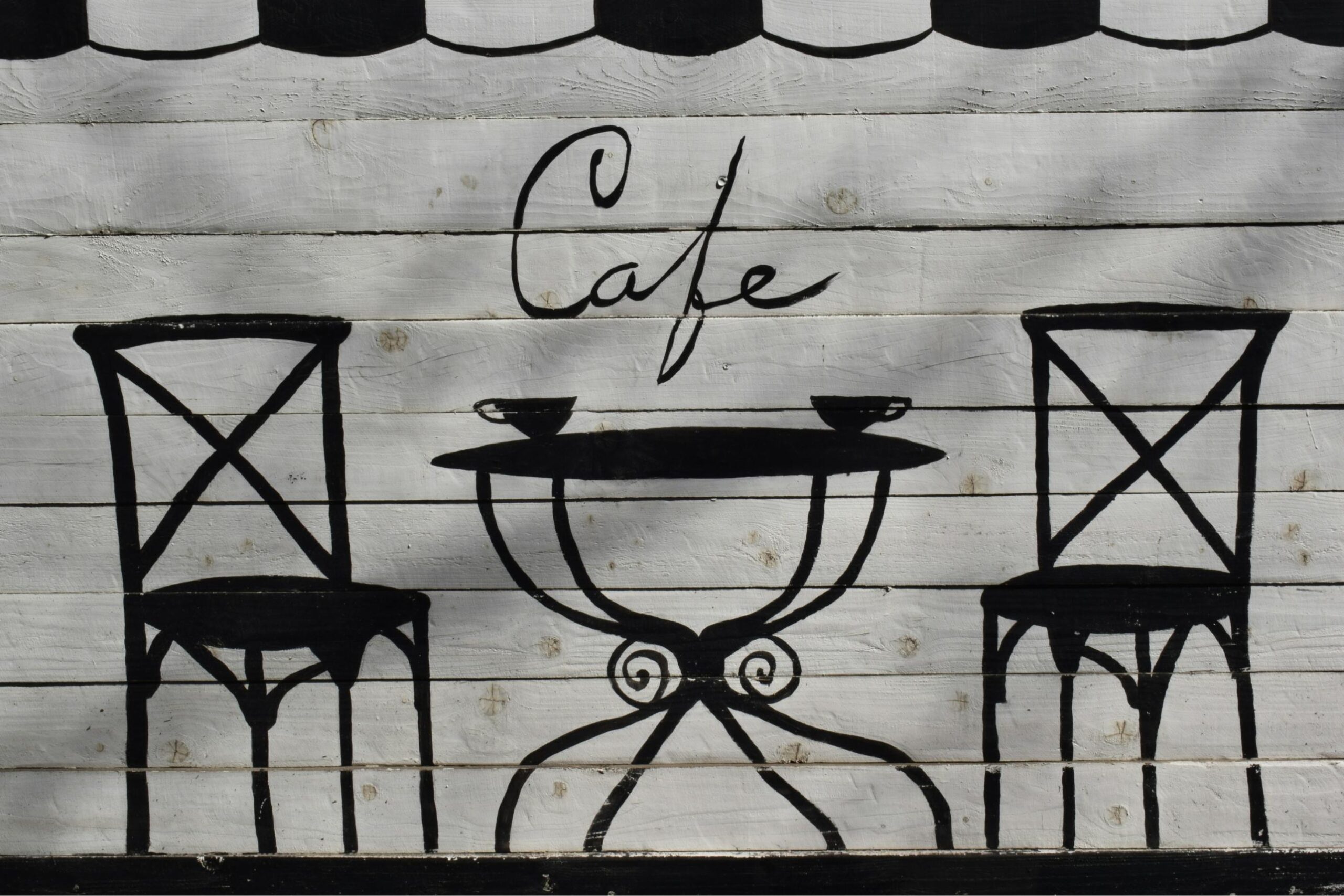 This image features a charming, hand-painted mural on a white wooden plank wall depicting a minimalist cafe scene. Two black bistro chairs flank a small table with two cups, set beneath elegant script that reads "Cafe”.