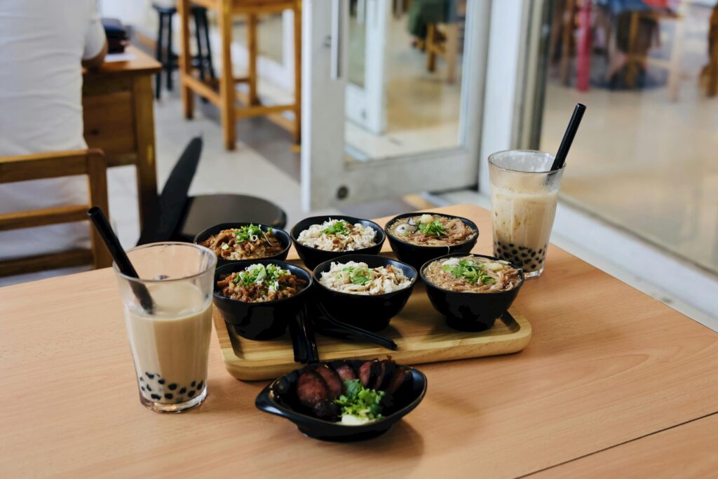 Six small black bowls of various Taiwanese dishes, including braised pork rice and noodles, are arranged on a wooden tray alongside two glasses of iced bubble tea. The meal is set on a light wood table in a brightly lit, modern cafe with a side dish of sliced sausage in the foreground.