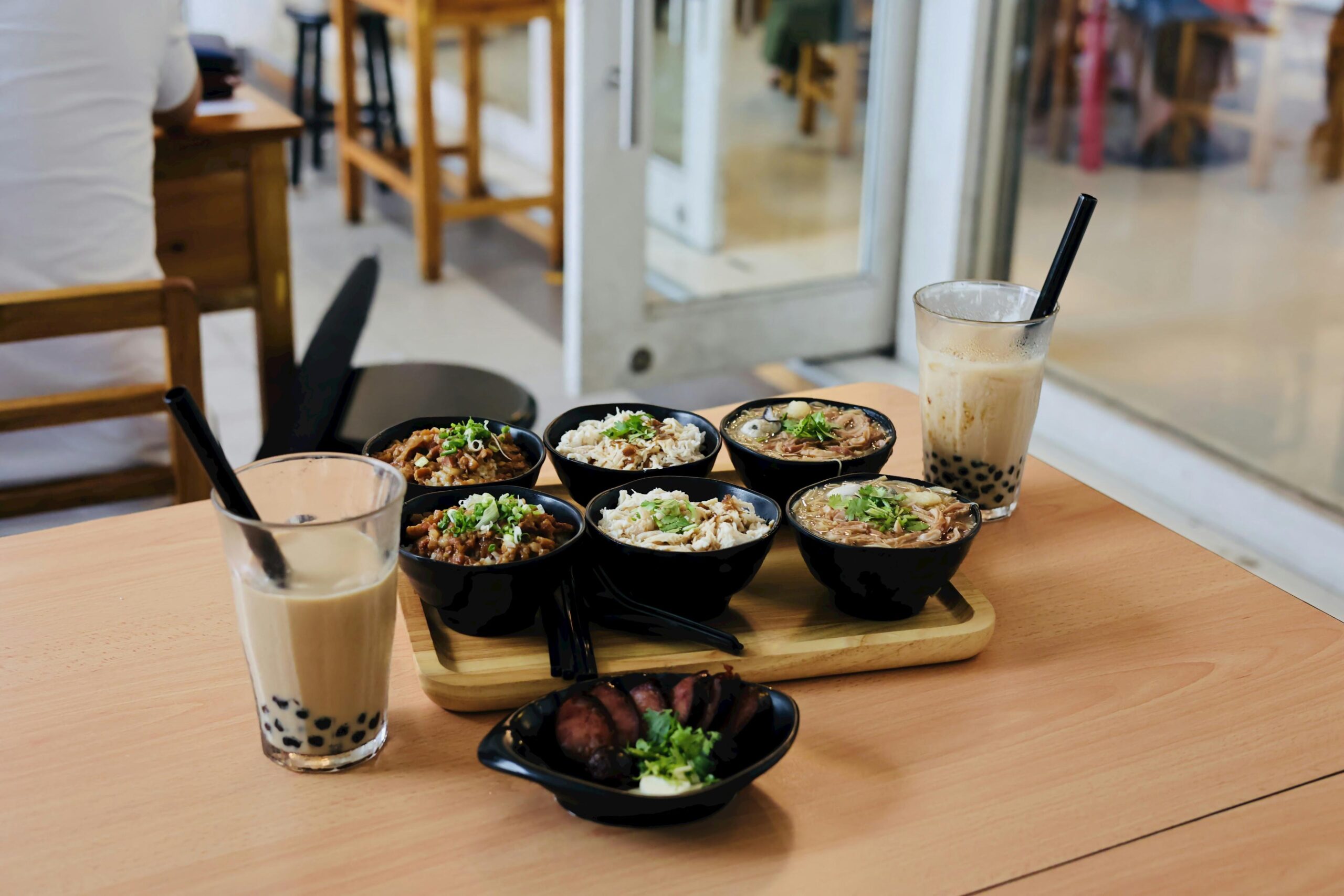 Six small black bowls of various Taiwanese dishes, including braised pork rice and noodles, are arranged on a wooden tray alongside two glasses of iced bubble tea. The meal is set on a light wood table in a brightly lit, modern cafe with a side dish of sliced sausage in the foreground.