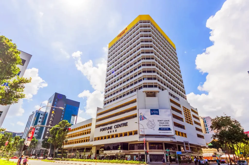 A low-angle exterior view of Fortune Centre building in Singapore under a bright, clear blue sky with fluffy white clouds. The white high-rise features distinct yellow architectural accents and large signage displayed on its facade.