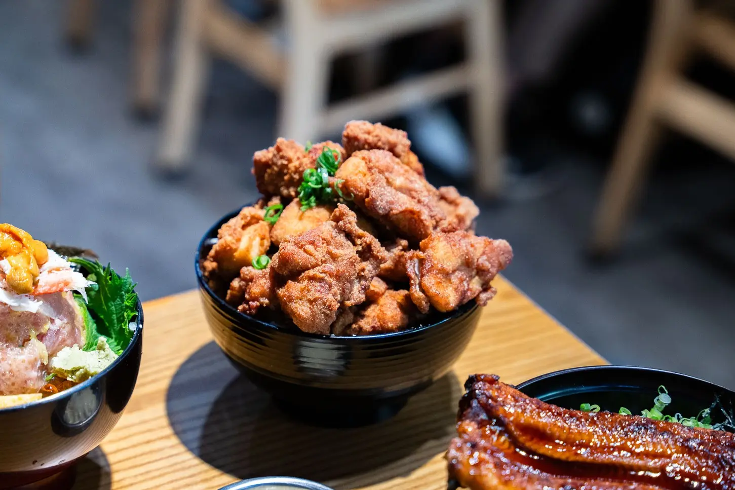 A high-angle, close-up shot shows a black bowl overflowing with a generous portion of golden-brown Japanese fried chicken (karaage), garnished with chopped green onions, sitting on a wooden table. To the left and right, parts of two other rice bowls are visible, one featuring seafood toppings and the other with a piece of grilled unagi eel.