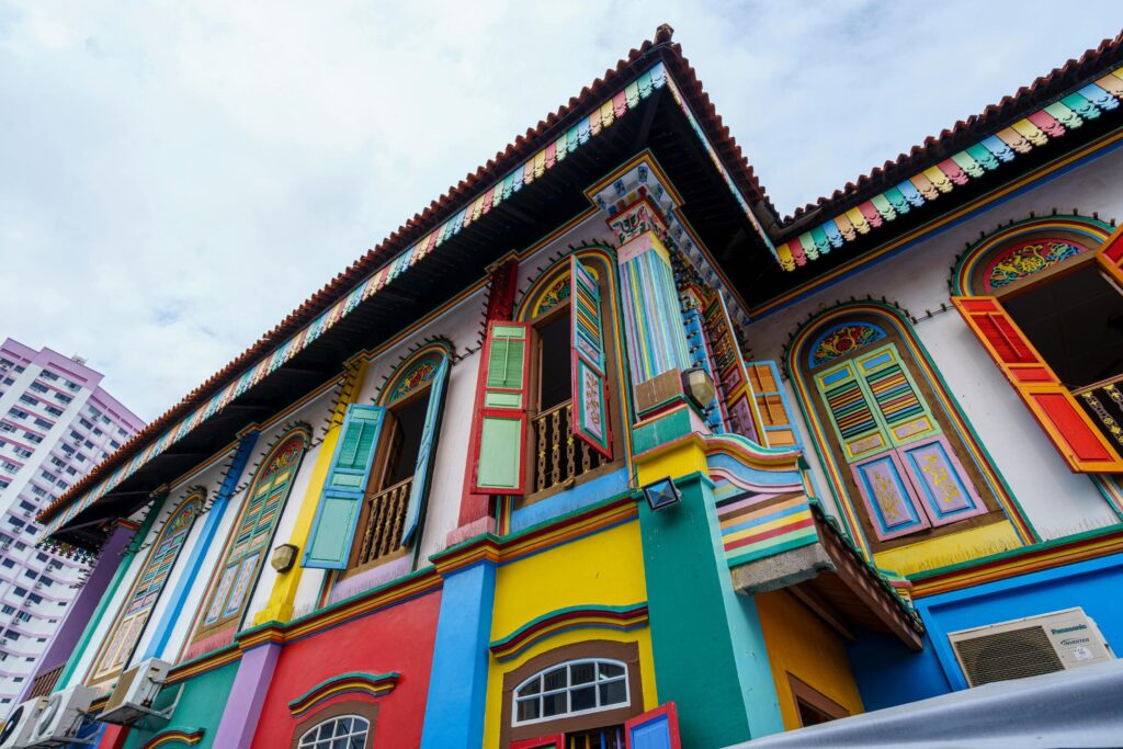 This vibrant image features a low-angle shot of the House of Tan Teng Niah in Little India, Singapore, showcasing its famous rainbow-colored facade and ornate architectural details. The building’s multi-colored shutters and intricately painted walls stand in bold contrast against a modern white high-rise and a soft, overcast sky.