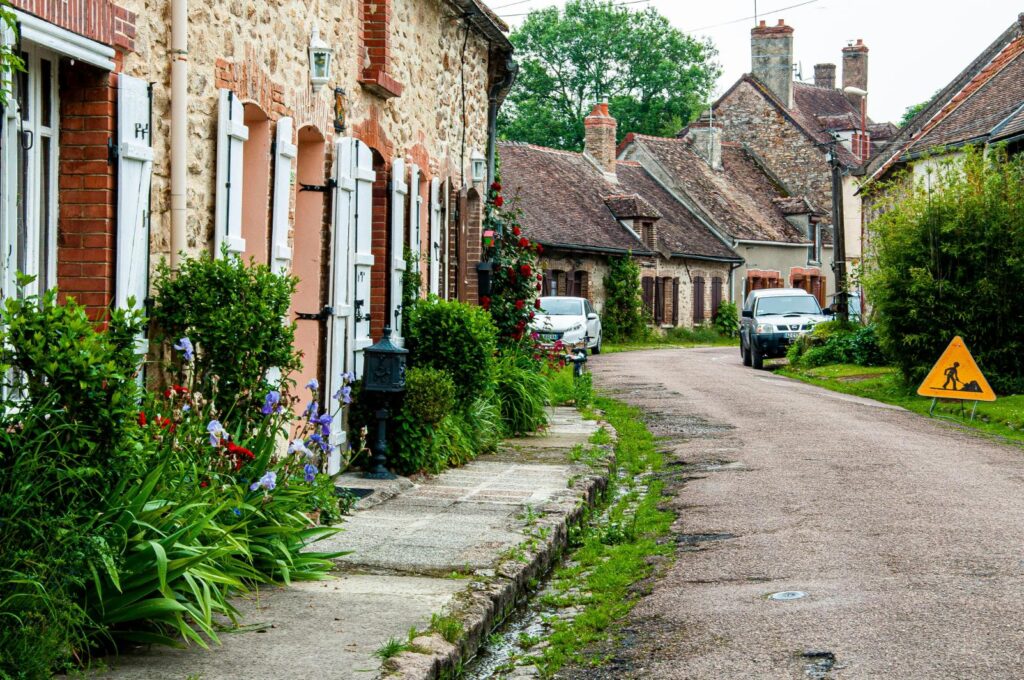 This charming street view showcases a row of rustic, stone cottages featuring white shutters and vibrant gardens filled with purple flowers and greenery. A narrow, winding road leads past parked cars and under a gray sky, capturing the quiet atmosphere of a European village.