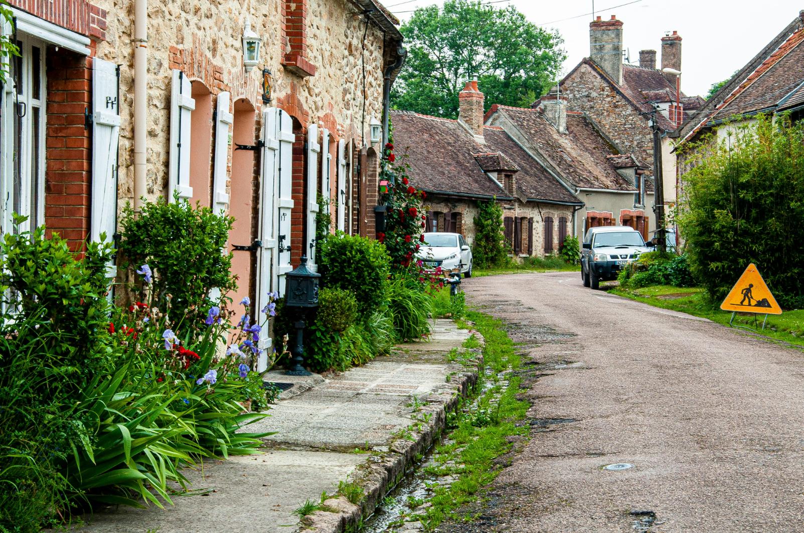 This charming street view showcases a row of rustic, stone cottages featuring white shutters and vibrant gardens filled with purple flowers and greenery. A narrow, winding road leads past parked cars and under a gray sky, capturing the quiet atmosphere of a European village.