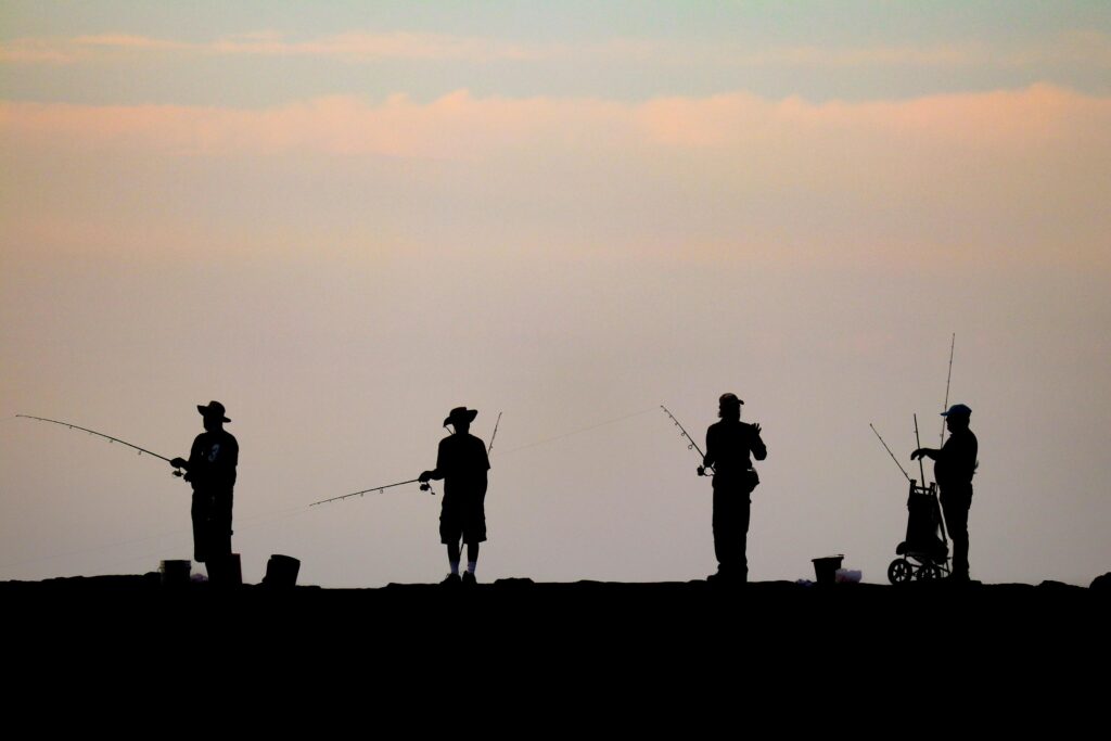 Four fishermen are captured in silhouette against a soft, pale sunset sky as they line a dark horizon. Each figure is spaced apart, holding a fishing rod and accompanied by gear like buckets and a small cart.