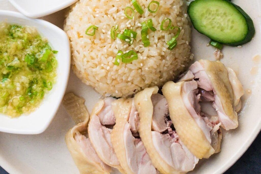 This plate features succulent slices of poached chicken served alongside a mound of seasoned rice topped with green onions and sesame seeds. The meal is accompanied by a fresh cucumber slice and a side of vibrant ginger-scallion dipping sauce.