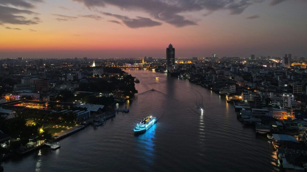A vibrant cruise ship illuminated with neon blue lights glides down a wide river at twilight, flanked by the dense cityscape and temples of Bangkok. The sky transitions from a deep orange glow on the horizon to soft purple clouds, reflecting beautifully off the dark water.