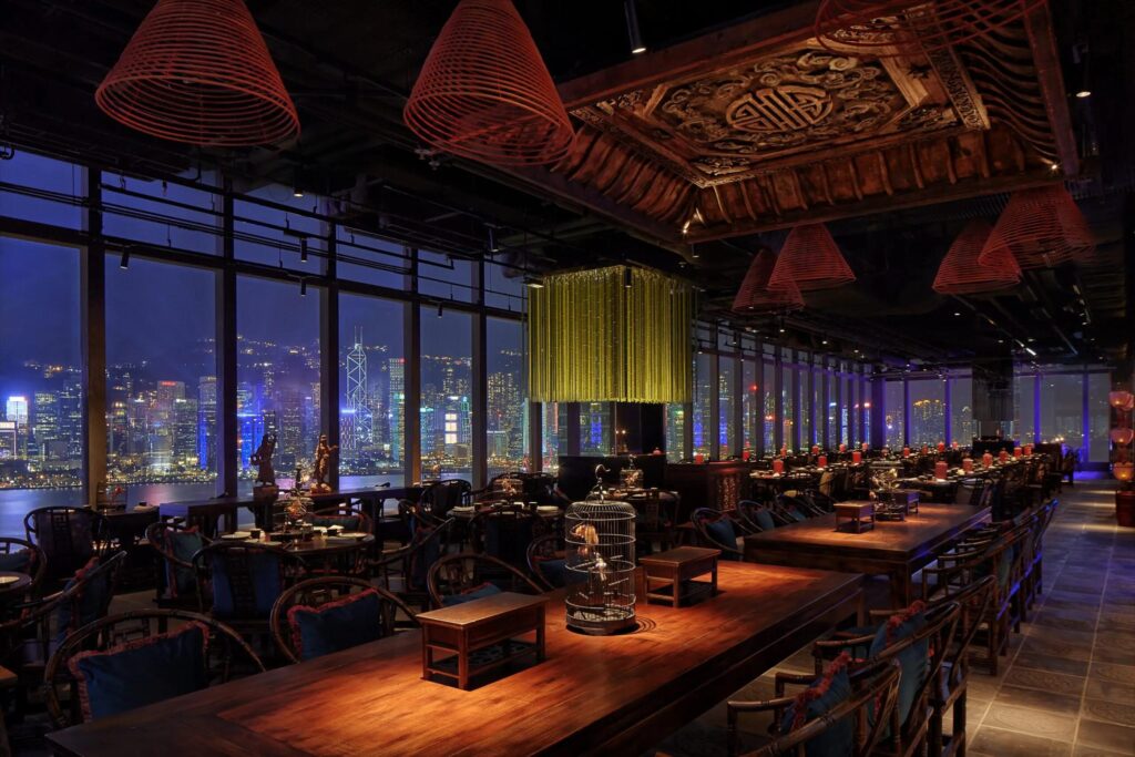 This upscale dining room features ornate traditional Chinese decor, including intricate wood carvings and birdcage accents, set against a stunning floor-to-ceiling view of a lit-up city skyline at night. The warm interior lighting and conical red lamps create an intimate atmosphere that contrasts beautifully with the cool, vibrant glow of the urban landscape outside.