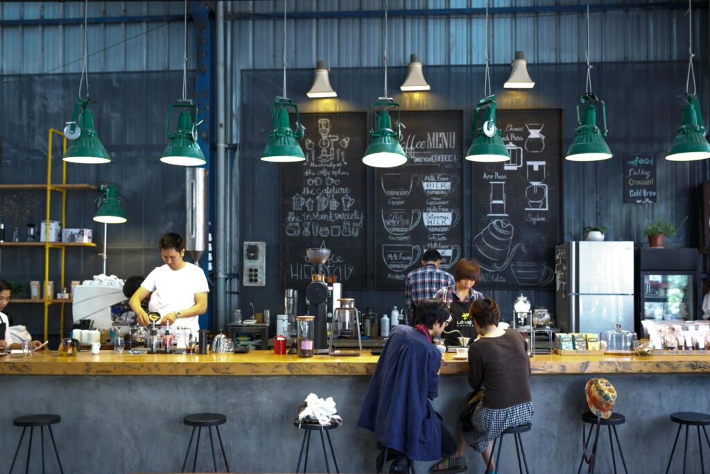 This industrial-style cafe features a long wooden counter where baristas prepare drinks beneath a row of vibrant green pendant lights. Behind the bar, large chalkboards display illustrated coffee menus and brewing methods against a corrugated metal wall.