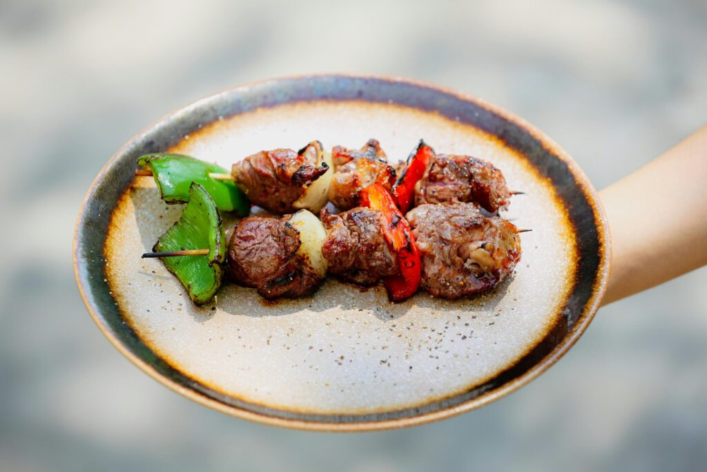 Two skewers of grilled meat and vegetables, including green bell peppers and onions, are presented on a rustic ceramic plate. An out-of-focus hand holds the plate outdoors, showcasing the charred edges and seasoning of the kebabs.