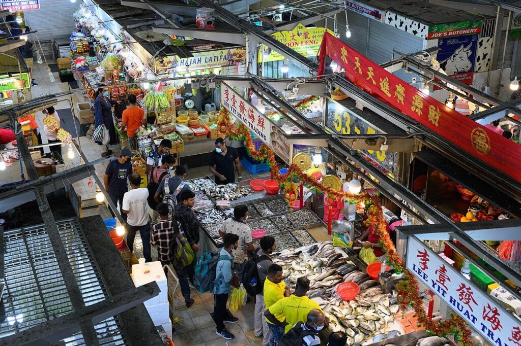This high-angle shot captures the bustling atmosphere of the Tekka Centre Wet Market, where shoppers gather around stalls overflowing with fresh seafood and local produce. The scene is filled with vibrant details, from the colorful shop banners and festive decorations to the diverse mix of people navigating the narrow, busy aisles.