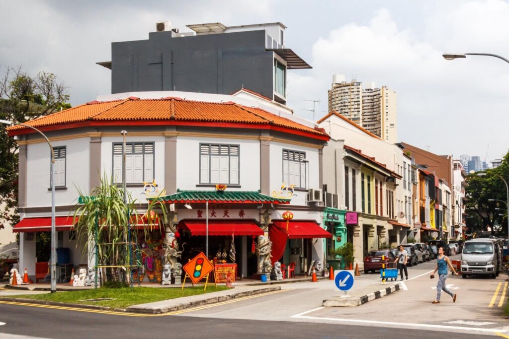 This street corner features a traditional white building with an orange-tiled roof and a Chinese temple storefront adorned with red awnings and decorative dragons. In the background, modern high-rise buildings and colorful shophouses line the road where pedestrians and vehicles are passing by.
