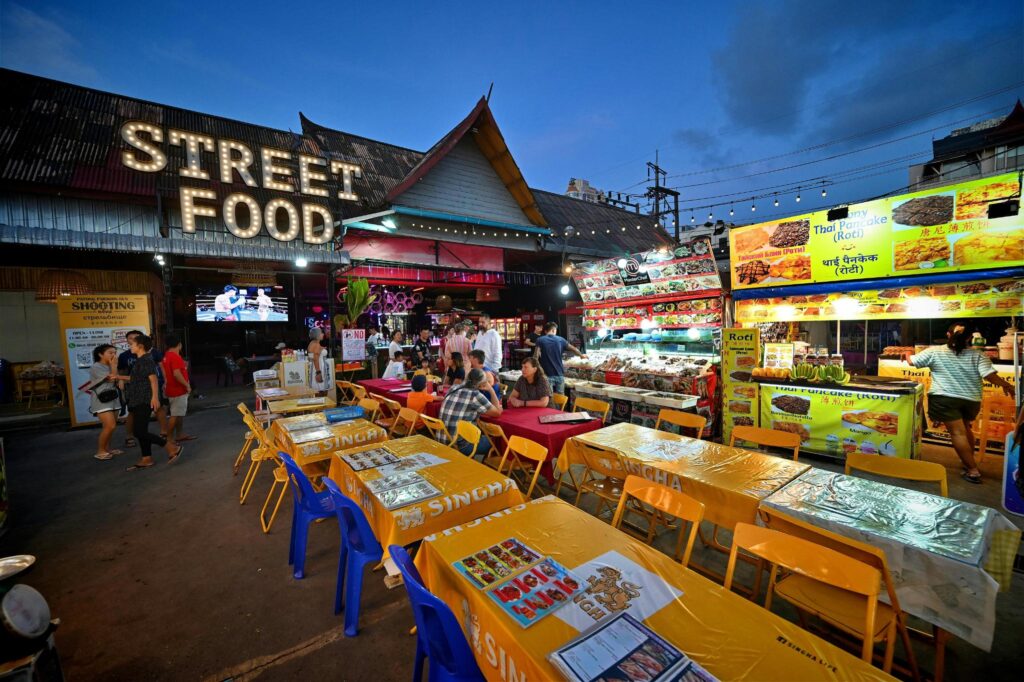 An outdoor night market features a "STREET FOOD" sign illuminated with light bulbs above a seating area with yellow-clothed tables and plastic chairs. Various stalls surround the dining area, including one specializing in "Thai Pancake (Roti)" and another displaying a wide array of seafood.