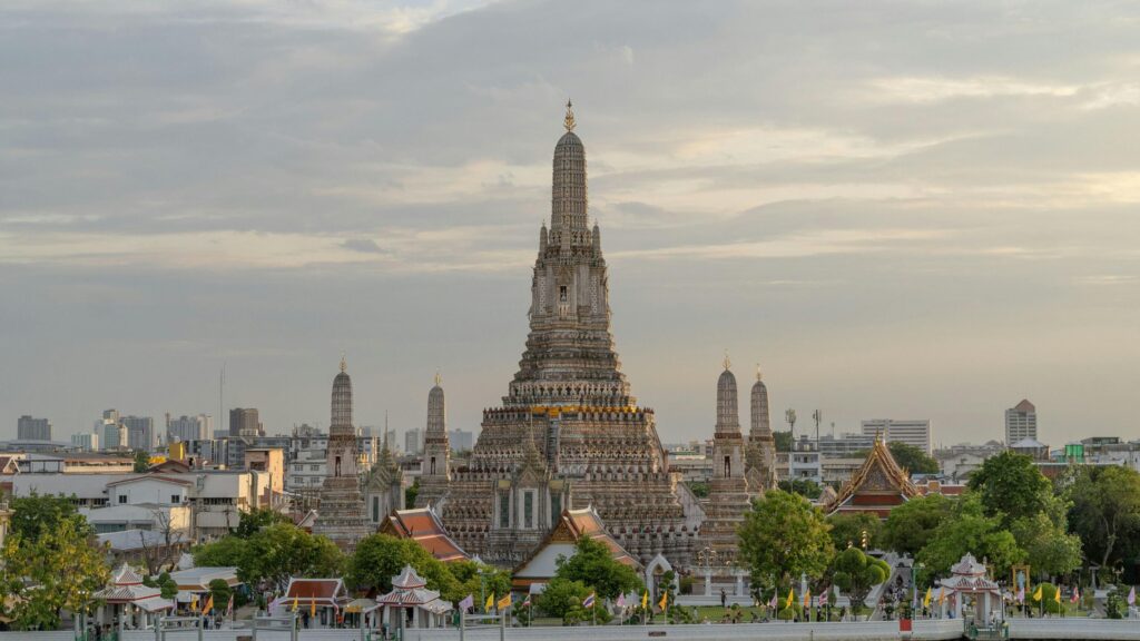 The majestic Wat Arun temple stands prominently against a cloudy sky, its intricate central spire surrounded by smaller prangs and lush greenery. Below the ornate structures, colorful flags line the riverfront as the city of Bangkok stretches into the distance.
