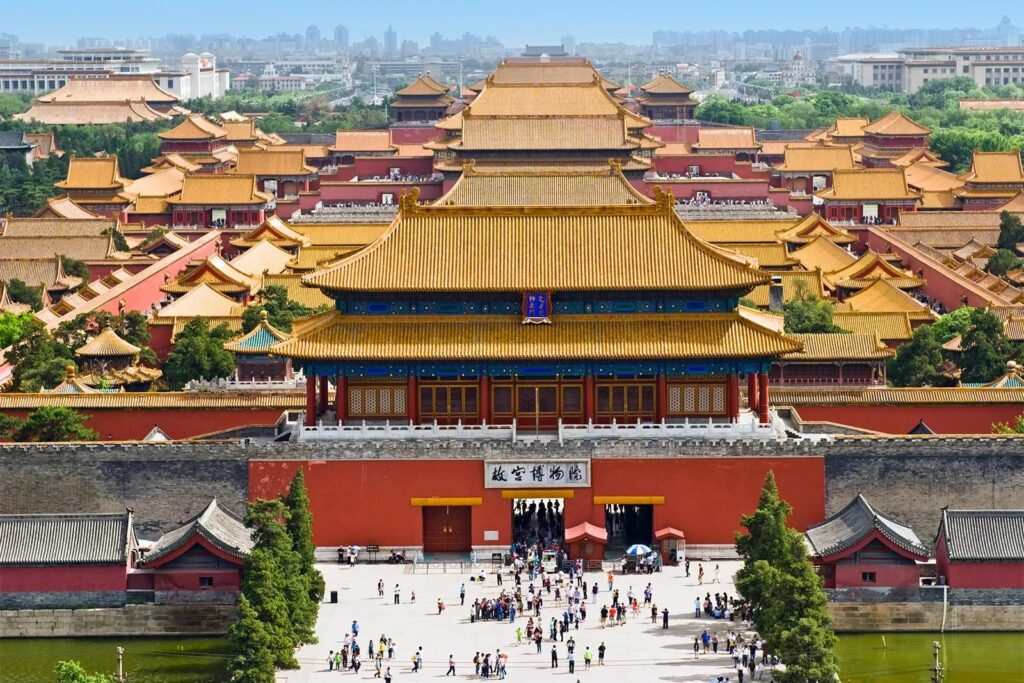 This wide-angle view showcases the Gate of Divine Might, the northern entrance to the Forbidden City in Beijing, with its iconic yellow-tiled roofs and red walls. Visitors are seen gathering at the entrance, while the vast complex of traditional imperial buildings stretches toward the modern city horizon.