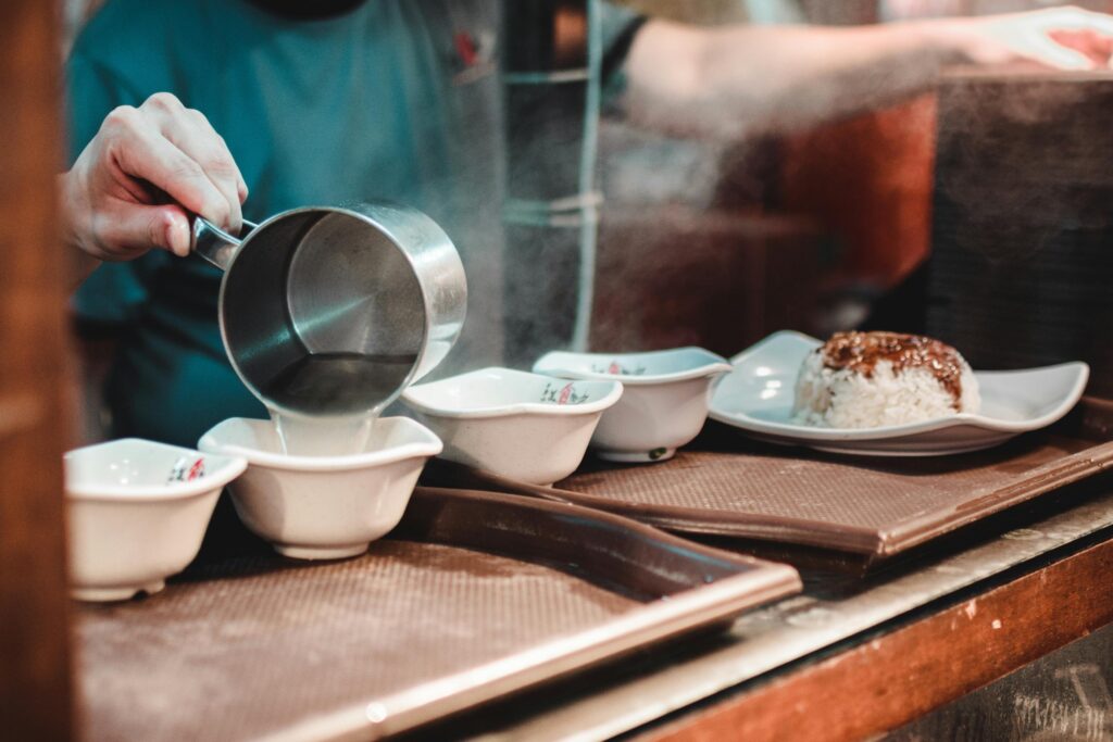 A cook pours steaming hot broth from a metal measuring cup into several small white bowls lined up on a serving tray. A finished plate of white rice topped with a dark sauce sits nearby, ready for service in a busy kitchen environment.