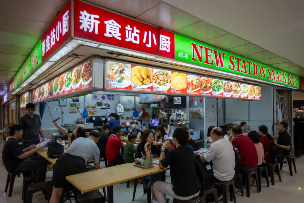 A brightly lit food stall named "New Station Snack Bar" is bustling with customers seated at long wooden tables in an indoor mall setting. Above the diners, a large red and green sign displays the shop's name in English and Chinese alongside vibrant photos of various menu items.