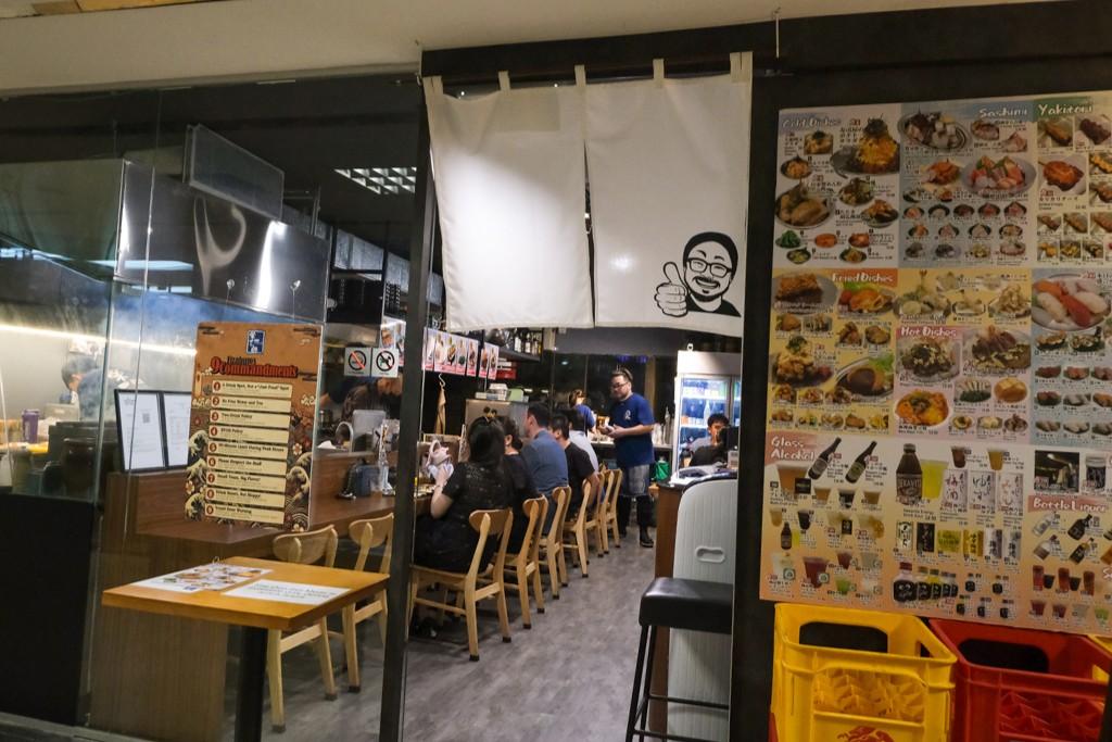 The image shows the entrance to Sakedokoro Eizaburo, a cozy Japanese eatery featuring a white noren curtain with a cheerful caricature. Patrons are seated along a narrow wooden counter inside, while an extensive illustrated menu of sashimi, yakitori, and drinks is displayed prominently on the exterior wall.