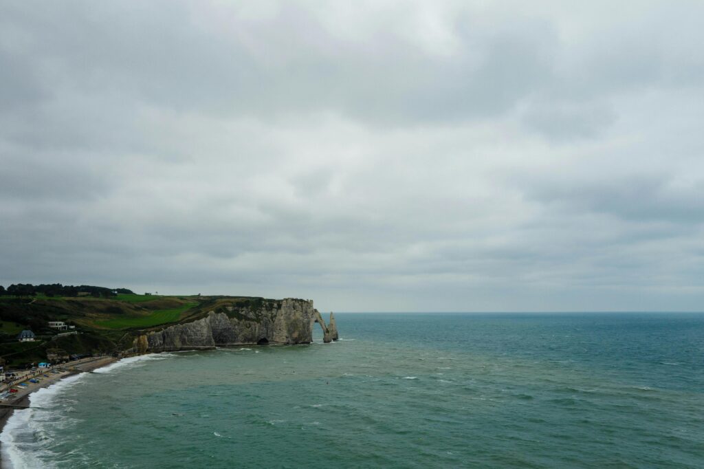 The image reveals a vast, moody seascape featuring the dramatic white chalk cliffs and natural rock arch of Étretat under a heavy, overcast sky. Below the grassy clifftops, a pebbled beach meets the churning turquoise waters of the English Channel, creating a somber yet majestic coastal atmosphere.