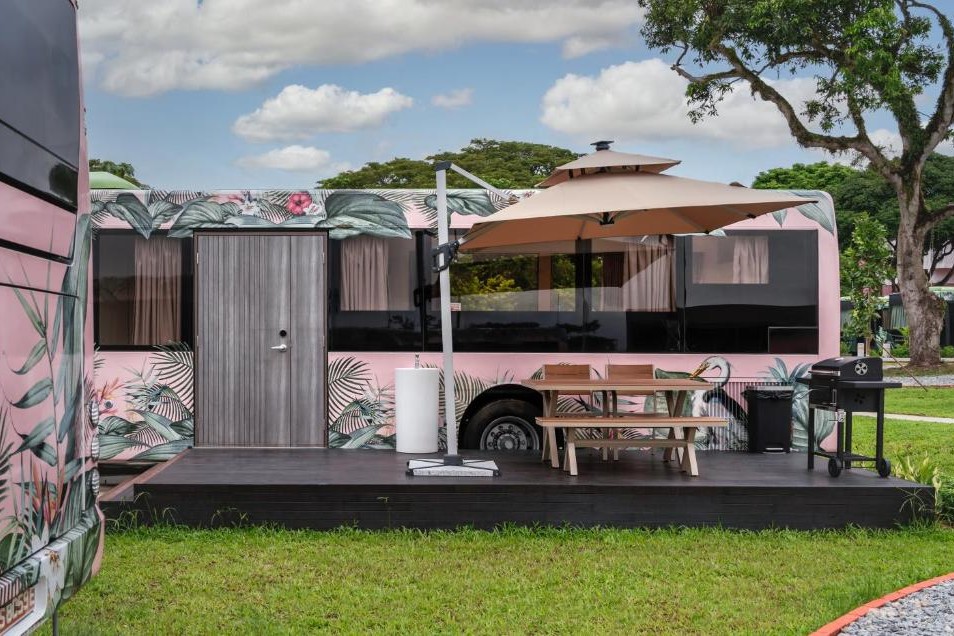 The image showcases a unique resort featuring a repurposed bus with a pink floral exterior and a private wooden deck equipped for outdoor dining. Inside, the bus is transformed into a modern living space with a tan sofa, sleek cabinetry, and large windows that provide a bright, airy feel.