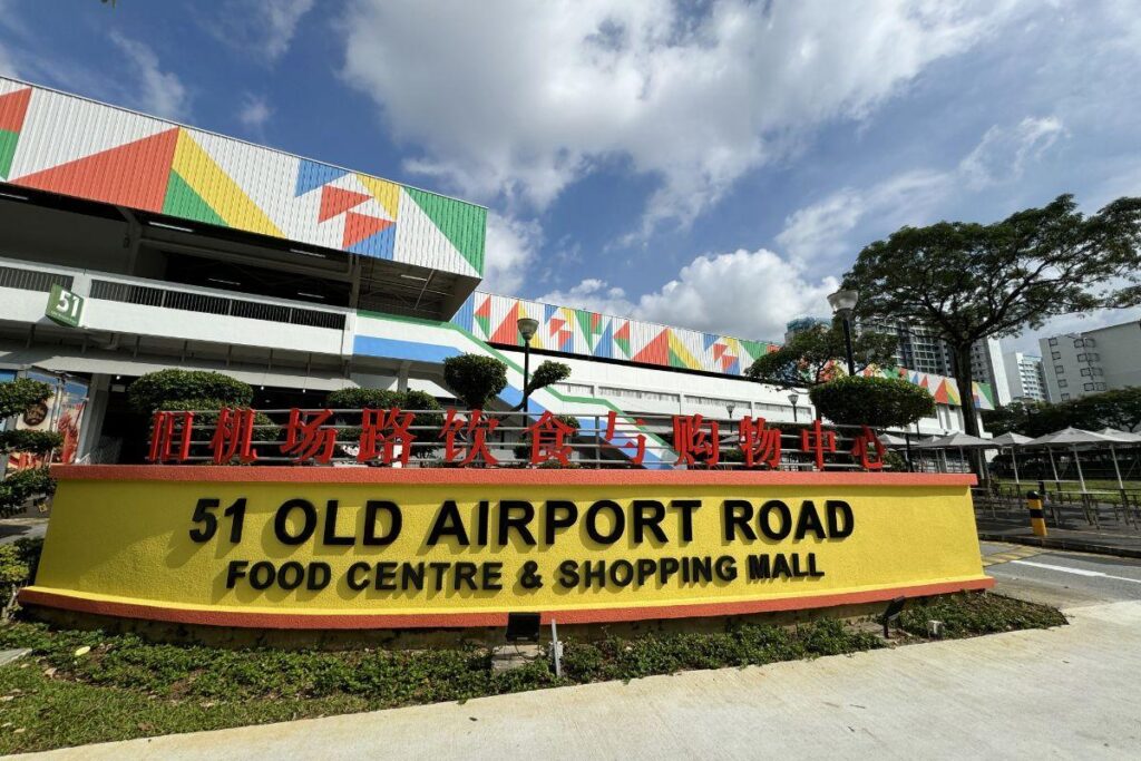A large yellow sign with red Chinese characters and black English text marks the entrance to the 51 Old Airport Road Food Centre & Shopping Mall. Behind the sign, a multi-story building features a white facade decorated with colorful, geometric triangular patterns under a bright, cloudy sky.
