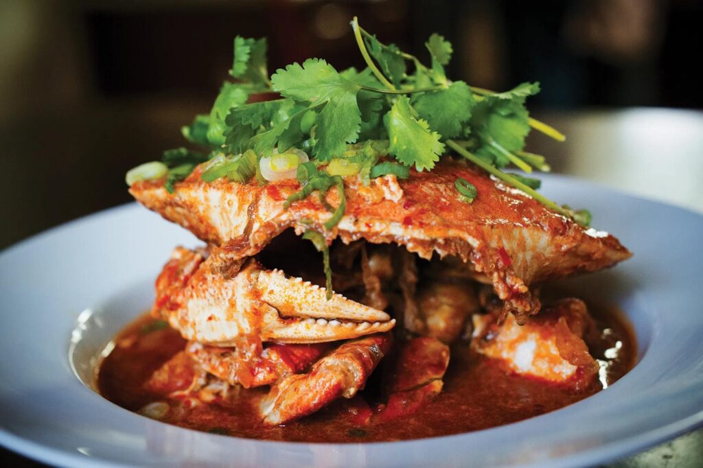 A whole crab is generously smothered in a thick, savory chili sauce and served in a shallow blue bowl. The dish is finished with a fresh garnish of chopped green onions and a large bunch of leafy cilantro on top.