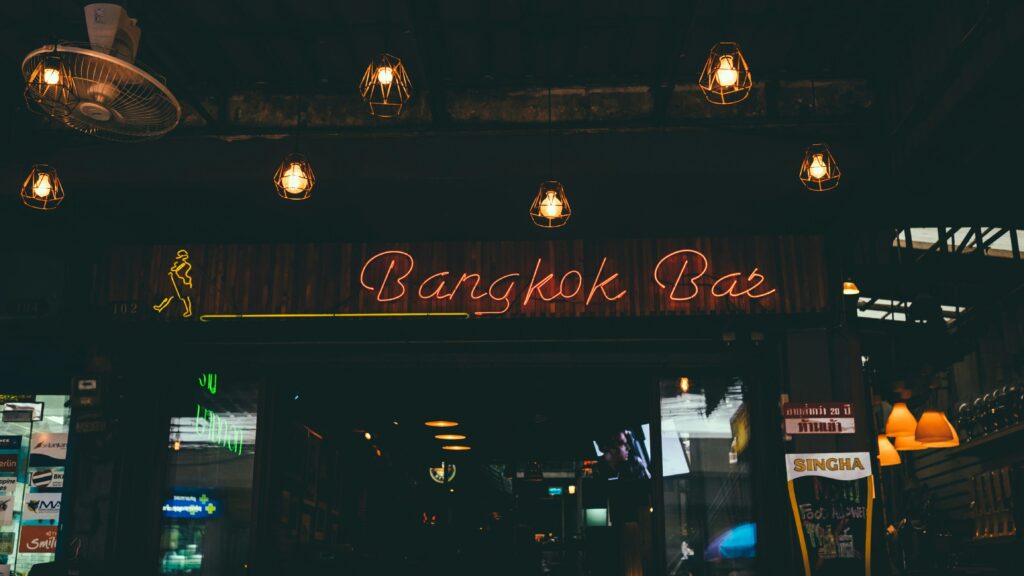 This image features a neon "Bangkok Bar" sign glowing in warm orange and red hues against a dark, industrial storefront. Geometric pendant lights hang from the ceiling, casting a soft amber glow over the entrance of this dimly lit nightlife spot.